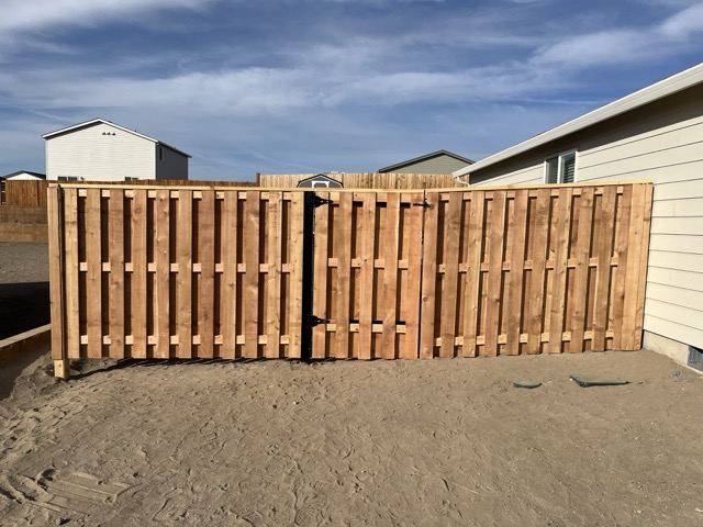 A newly built wooden privacy fence with a double gate in the center, set on sandy ground with houses and blue sky in the background.